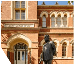 A closely cropped image of a part of the front of a school building, where you can see the gothic style of the window and door frames. A statue of a suited Bastow, refined with one arm behind his back, is in the foreground.