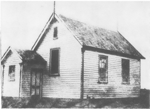An extremely old looking black and white photo of a non-descript rural school building. A small simple wooden structure with a gabled roof.
