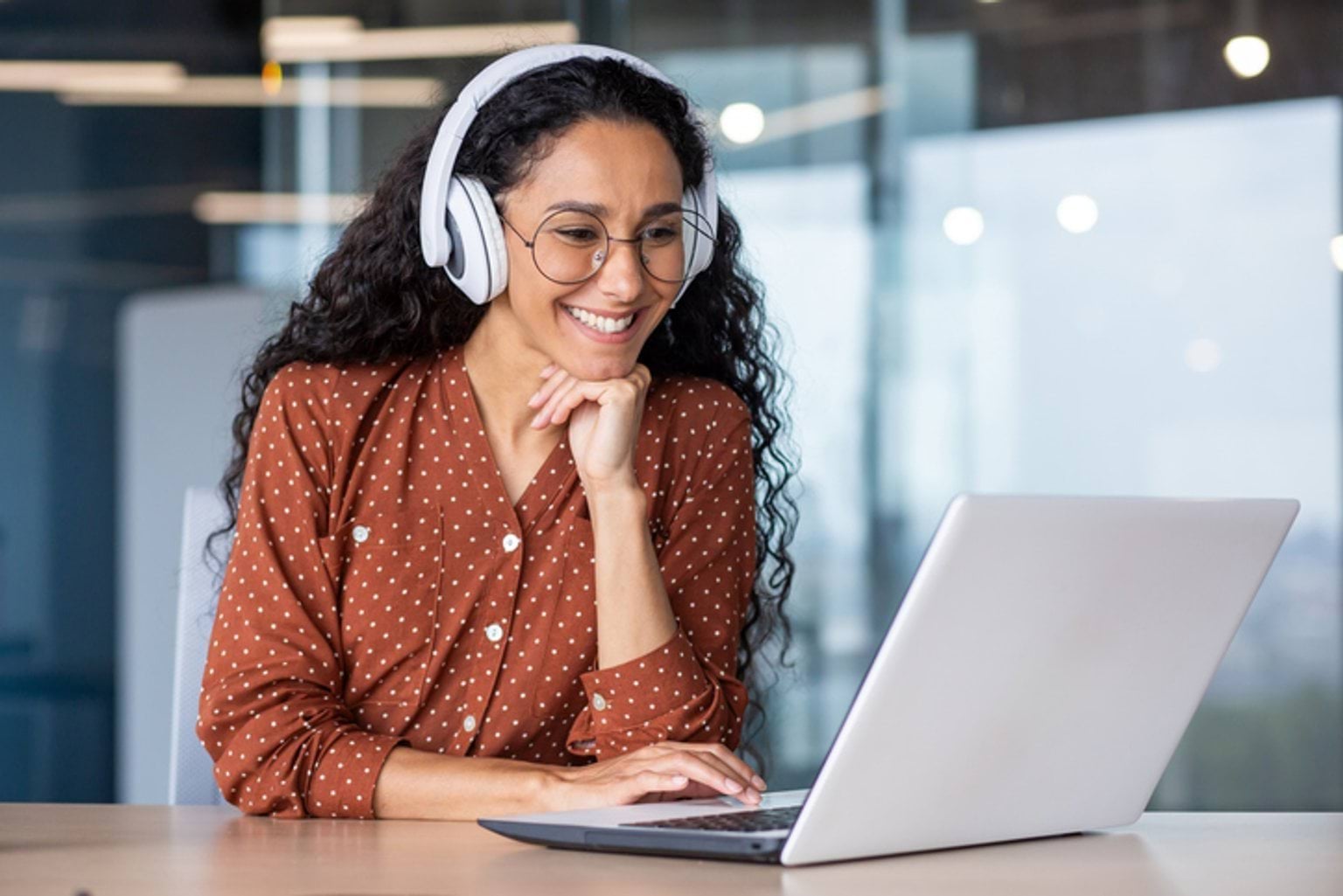 A stock image of a young woman looking at her laptop. She is smiling and has headphones on.