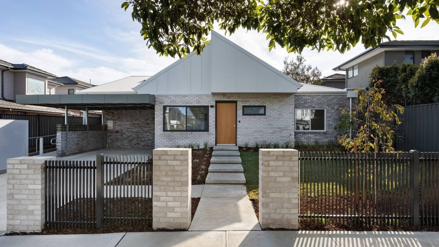 Modern grey brick house with a triangular roof, framed by green foliage. It features a wooden door, large windows and a path leading to the entrance.