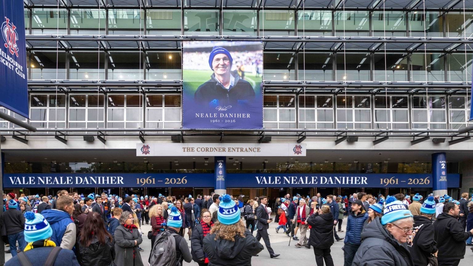 Crowds wearing FightMND beanies gather outside the MCG for Neale Daniher AO's State Funeral Service. There is a large image of Neale on the outside of the stadium.