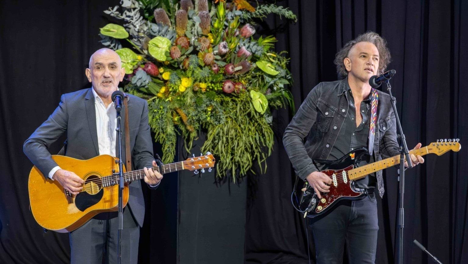 Musician Paul Kelly performs Leaps and Bounds at Neale Daniher AO's State Funeral Service. He is playing guitar and has another guitarist next to him.