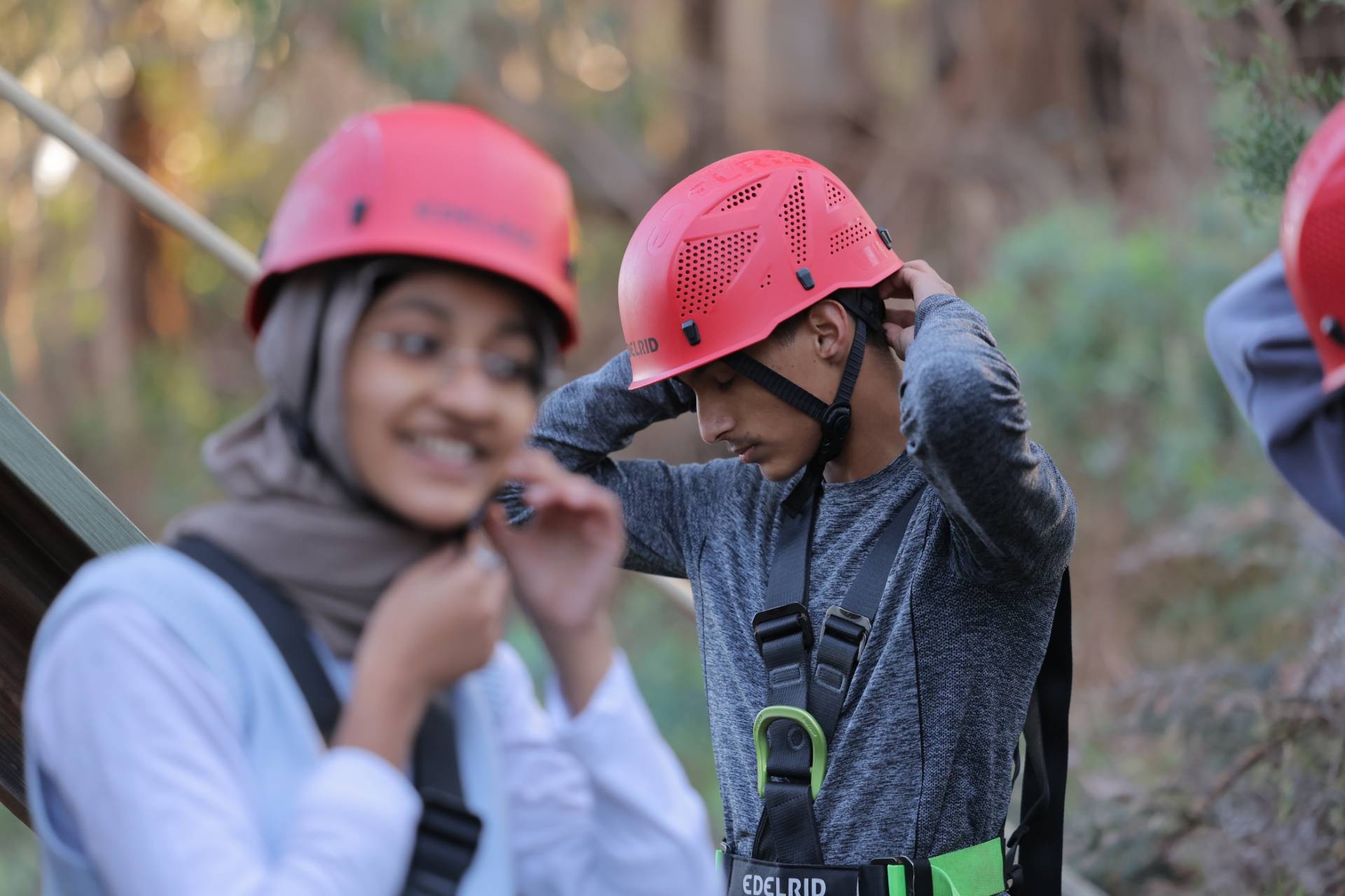 Three people wearing pink safety helmets and climbing harnesses in a wooded outdoor setting; the person in the center adjusts their helmet straps while another person in the foreground smiles and fastens their helmet, with blurred trees in the background.