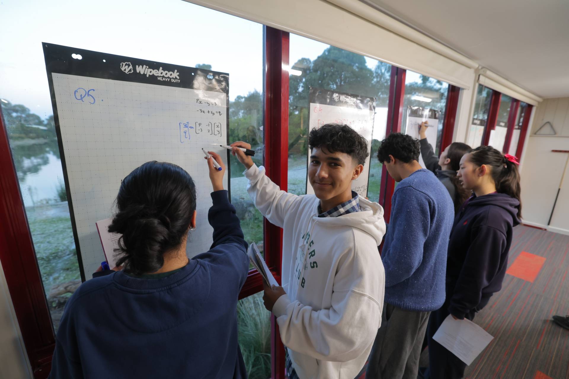 A group of students collaborating on a math activity inside a classroom, writing equations and notes on large whiteboards near windows. One student in a white hoodie looks toward the camera while others work together on problems in the background.