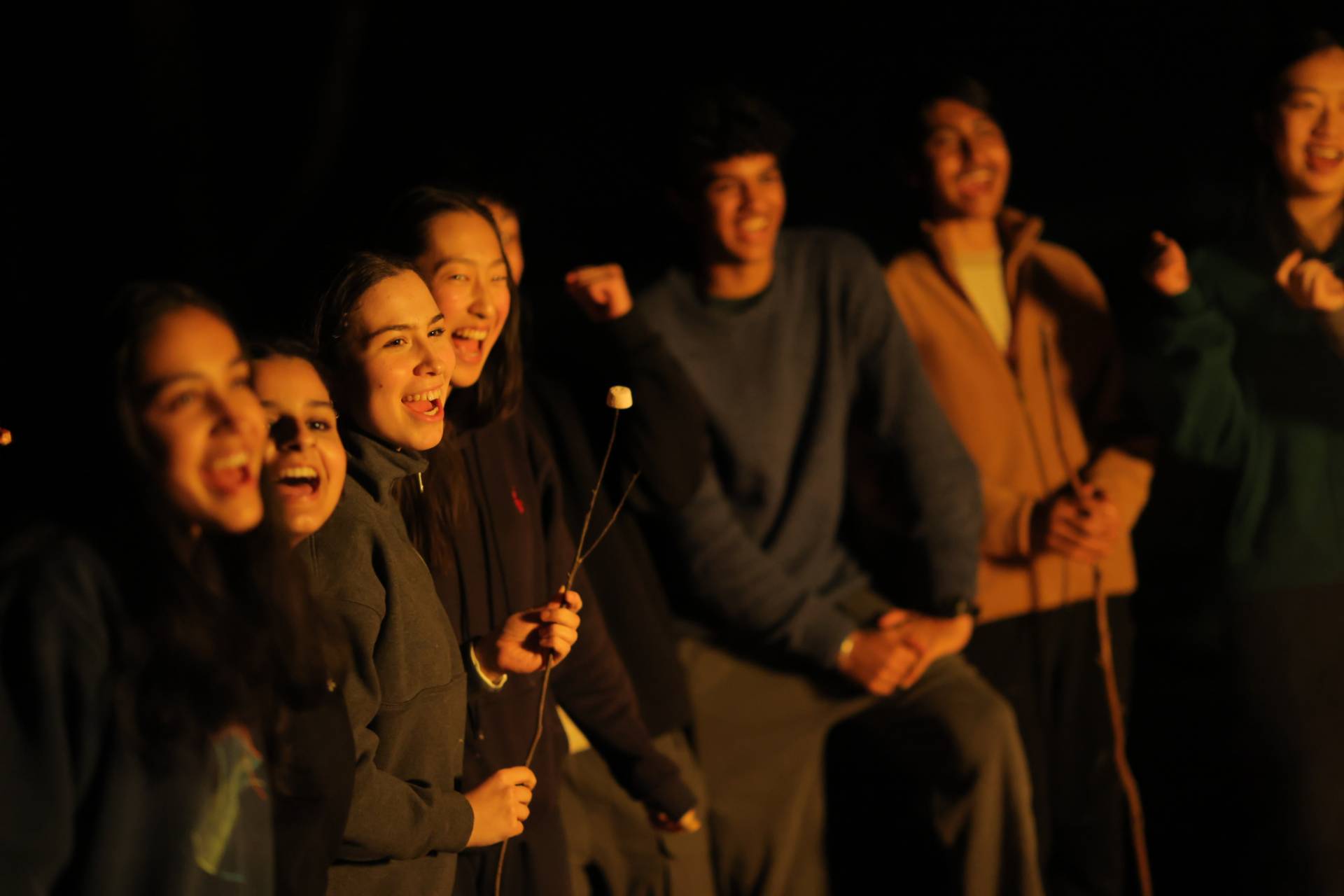 A group of people gathered in a dark outdoor setting, illuminated by warm firelight, smiling and laughing together while holding long sticks with marshmallows or similar items, creating a cozy campfire atmosphere.