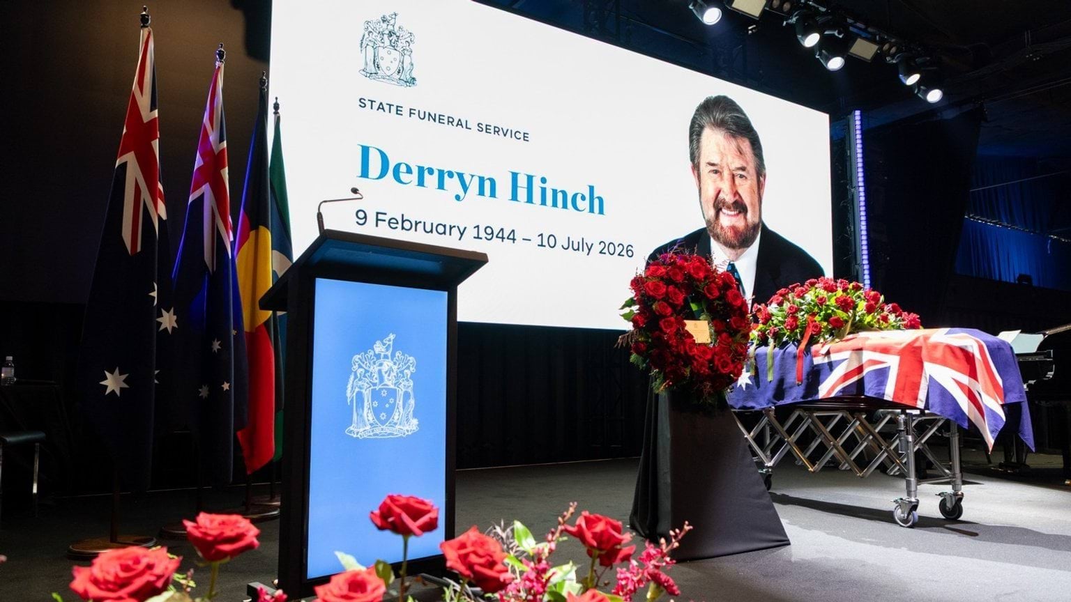 Image shows the stage for Mr Derryn Hinch's State Funeral Service, including his image on screen, a wreath, a lectern and his coffin