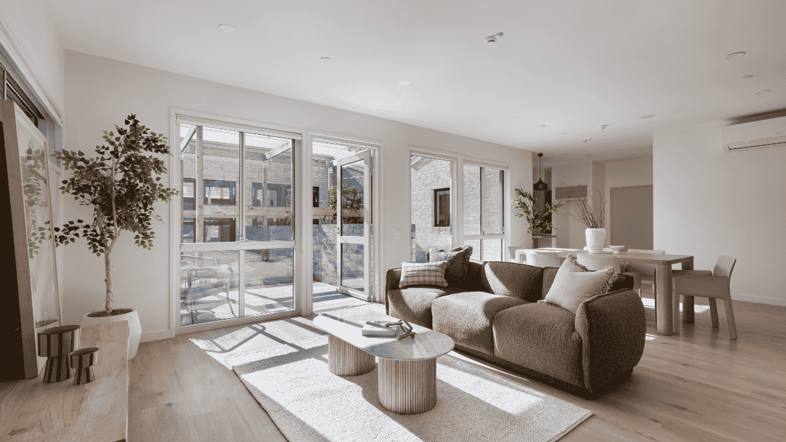 A modern living room with a large window, a brown couch, a small round coffee table, some greenery and a dining table in the background.