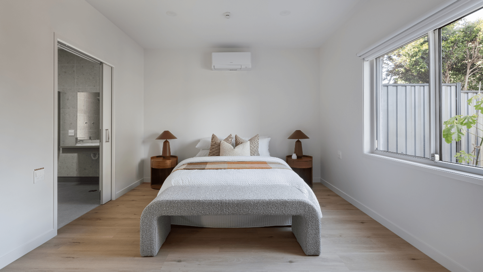 A modern bedroom with a light-coloured bed frame, neutral bedding and two bedside tables with lamps, featuring a large window letting in natural light and a sliding door into an ensuite bathroom..