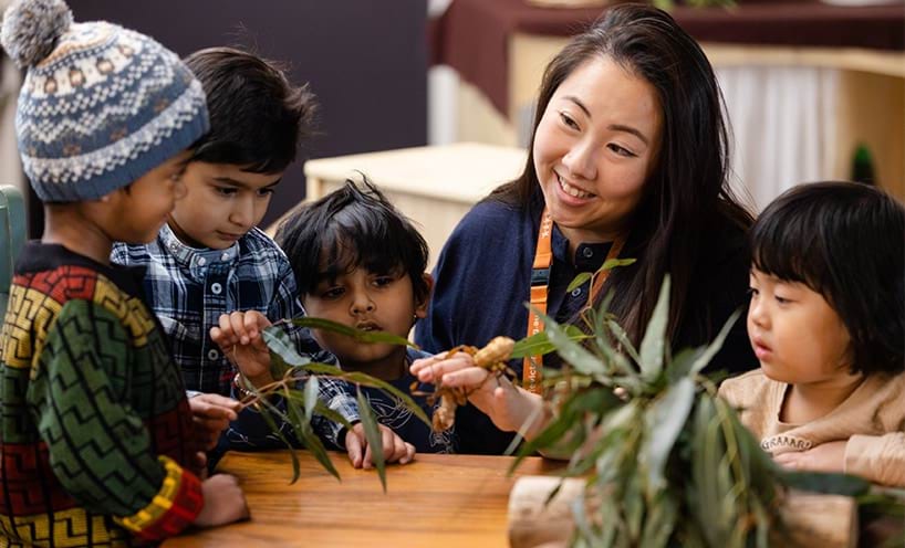 An educator interacting with children holding gum leaves around a table