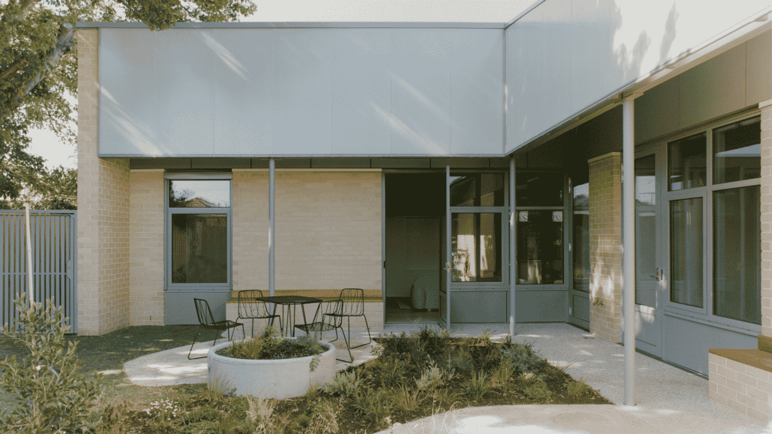 A modern building exterior with a patio area featuring a table and chairs, surrounded by plants and greenery.