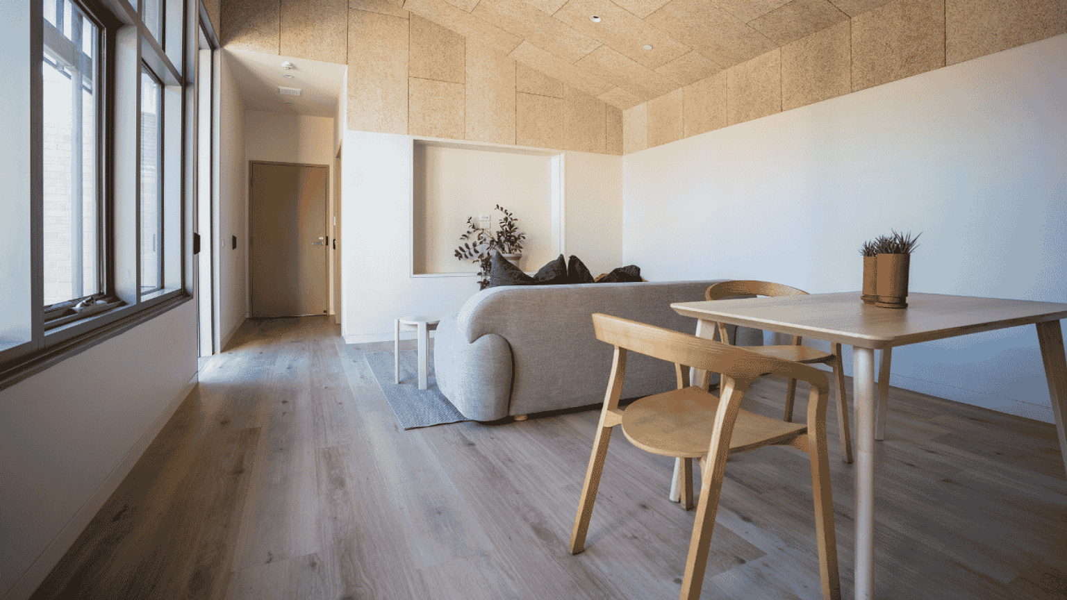 A modern and minimalistic interior design featuring a light wood dining table and chair, a cream couch and large windows that allow natural light into the space.