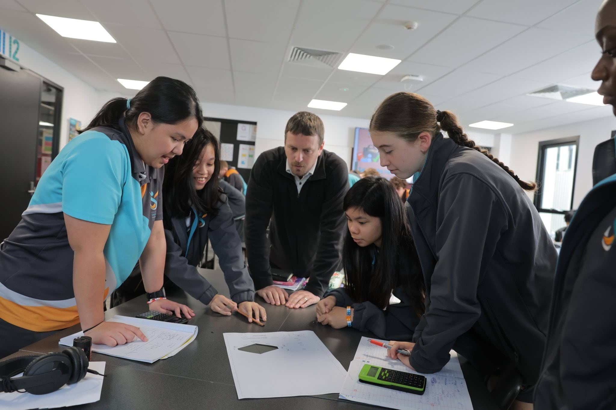 A group of students working at a table supervised by a teacher. The students are focused on calculations and planning.