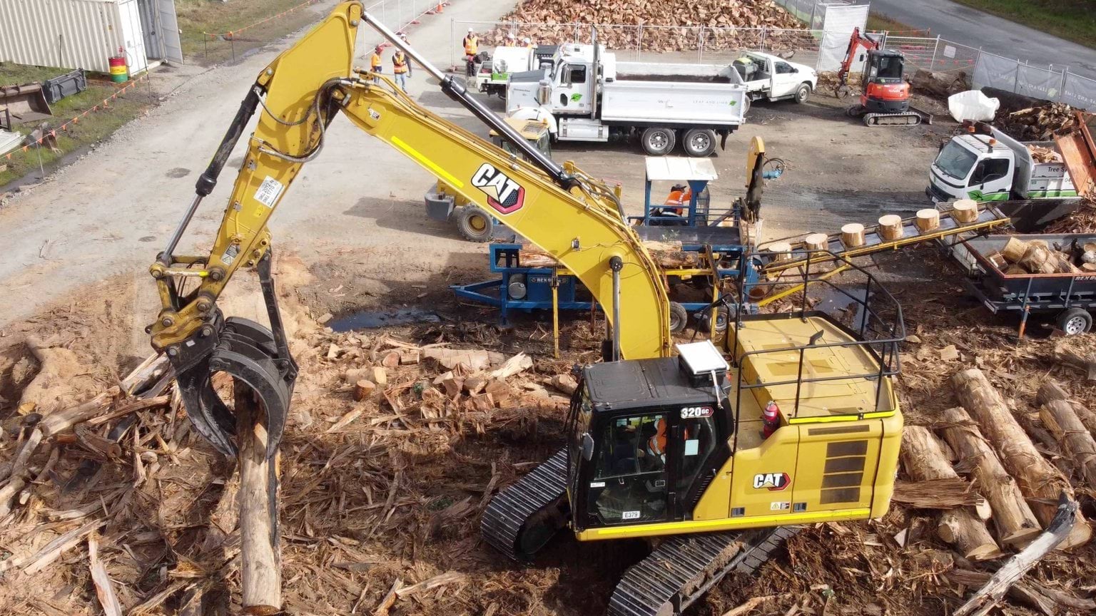 A yellow material handline machine uses a grabber to pick up logs from a pile of wood. There are trucks and chopped logs in the background.