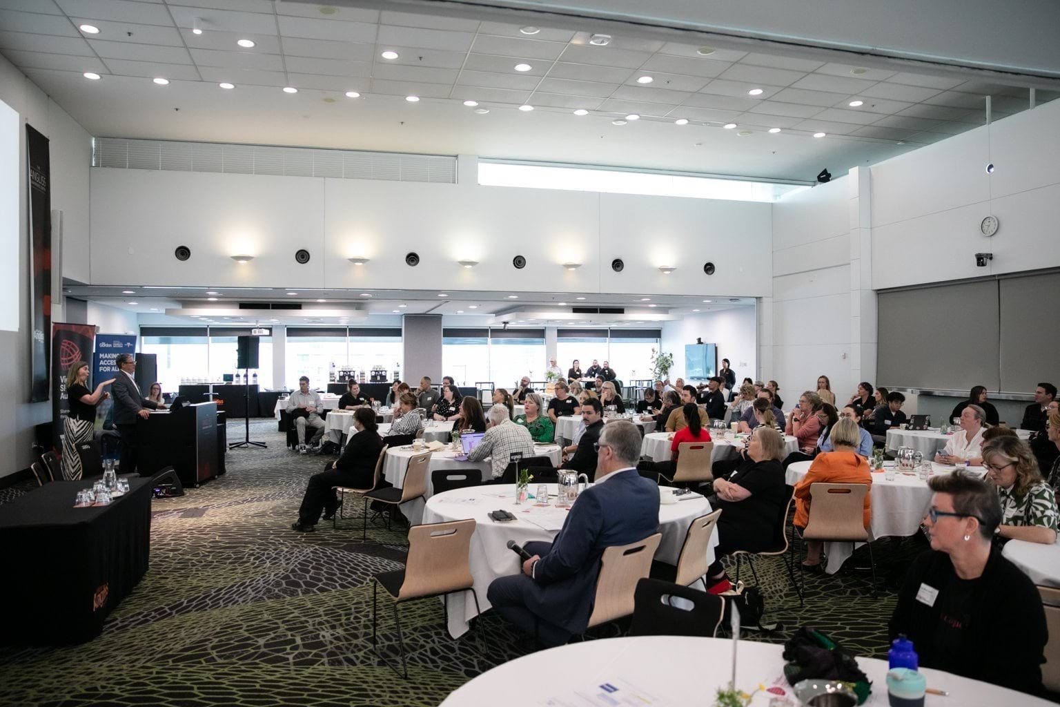 People sitting in an event conference space listening to the speakers facing the front of the room 