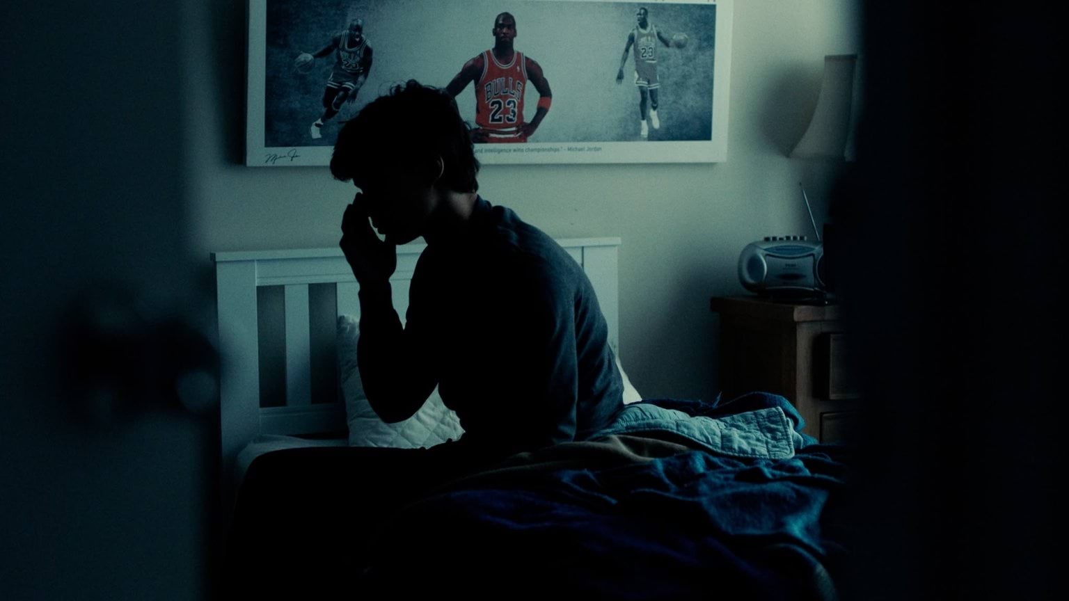 A shadowed room, dark lighting shows a young teenage male sitting at the edge of his bed rubbing his forehead in fatigue.