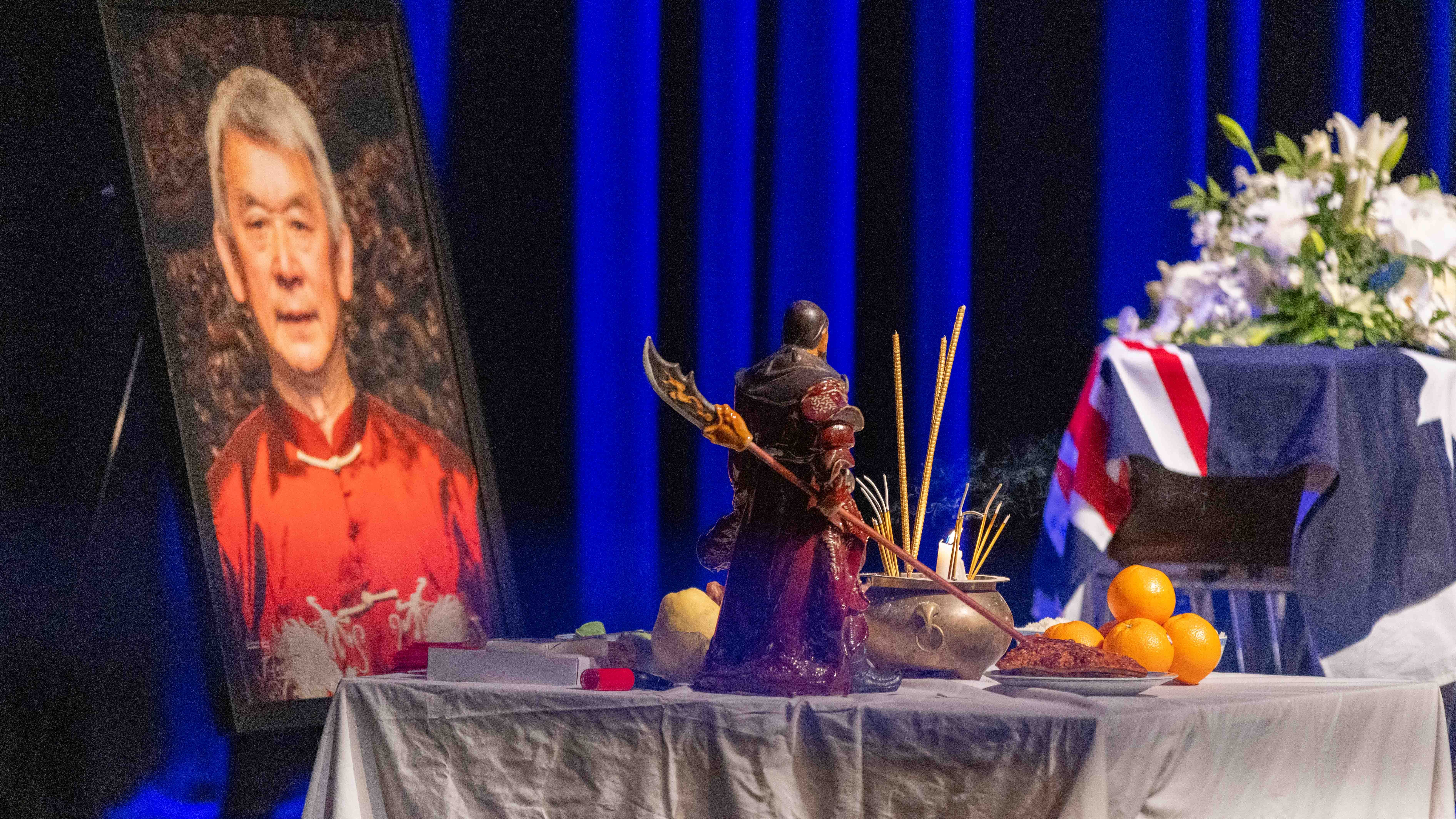 Image shows an altar on the stage at Russell Goldfield Jack AM's State Funeral Service. There is a figurine, fruit and joss sticks on the altar. Behind the altar is a portrait of Russell Jack and his coffin
