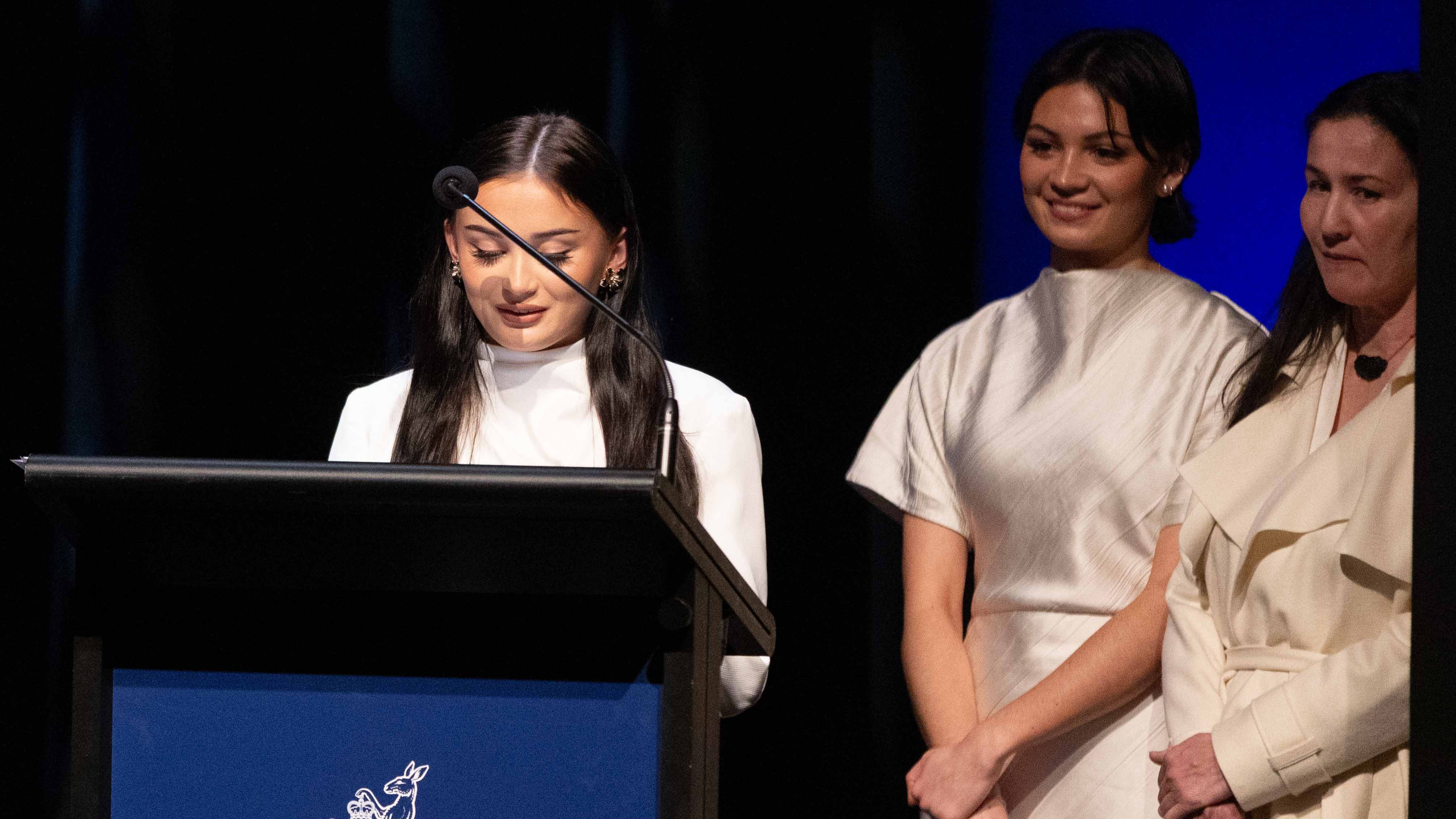 Amelita speaks at a lecturn to pay tribute to her grandfather Russell Goldfield Jack AM. Her sister and mother are standing behind her.