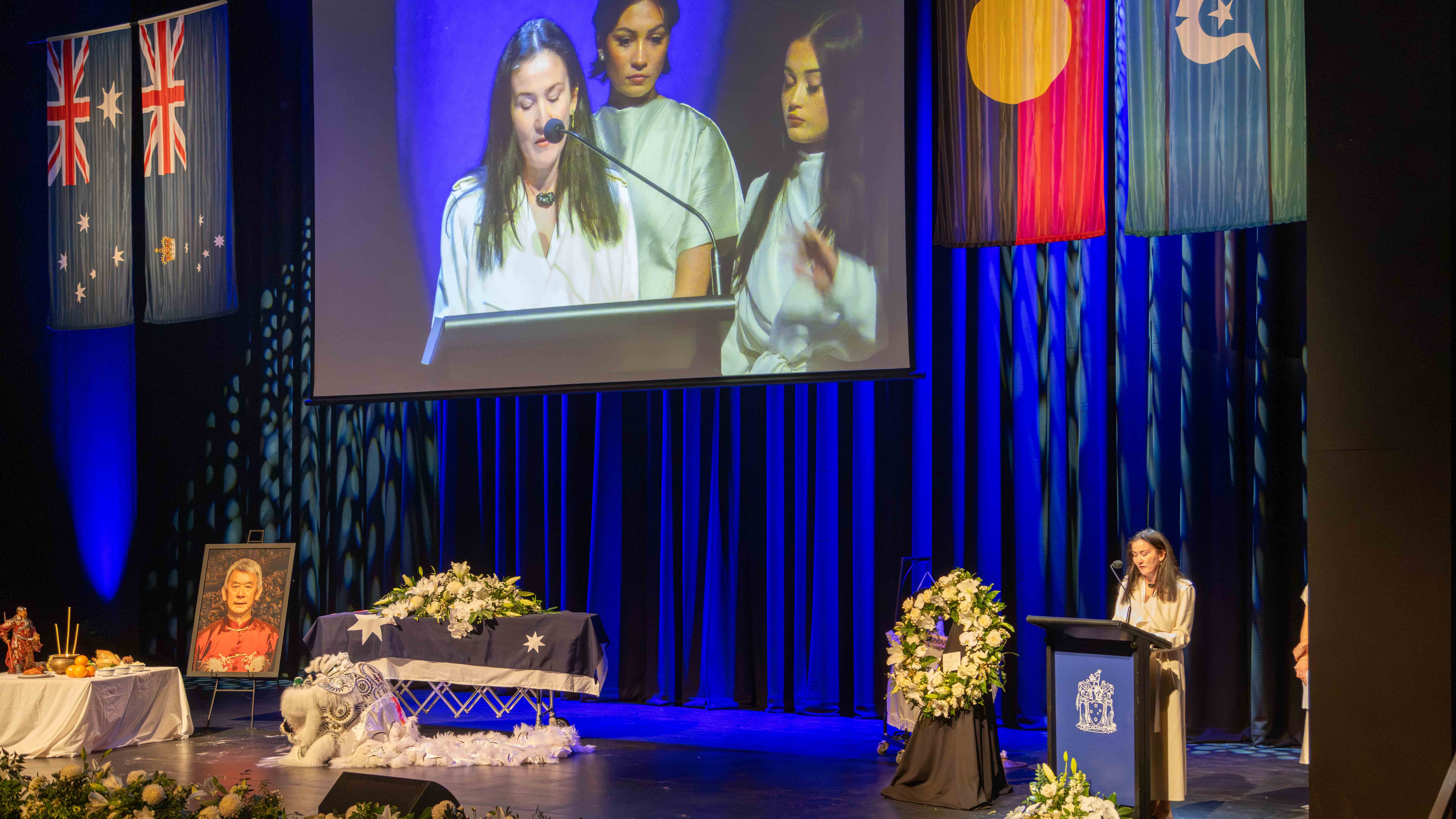 Anita Jack speaks from a lectern at the State Funeral Service of her father. On stage is Russel Goldfield Jack AM's coffin and a portrait of him.