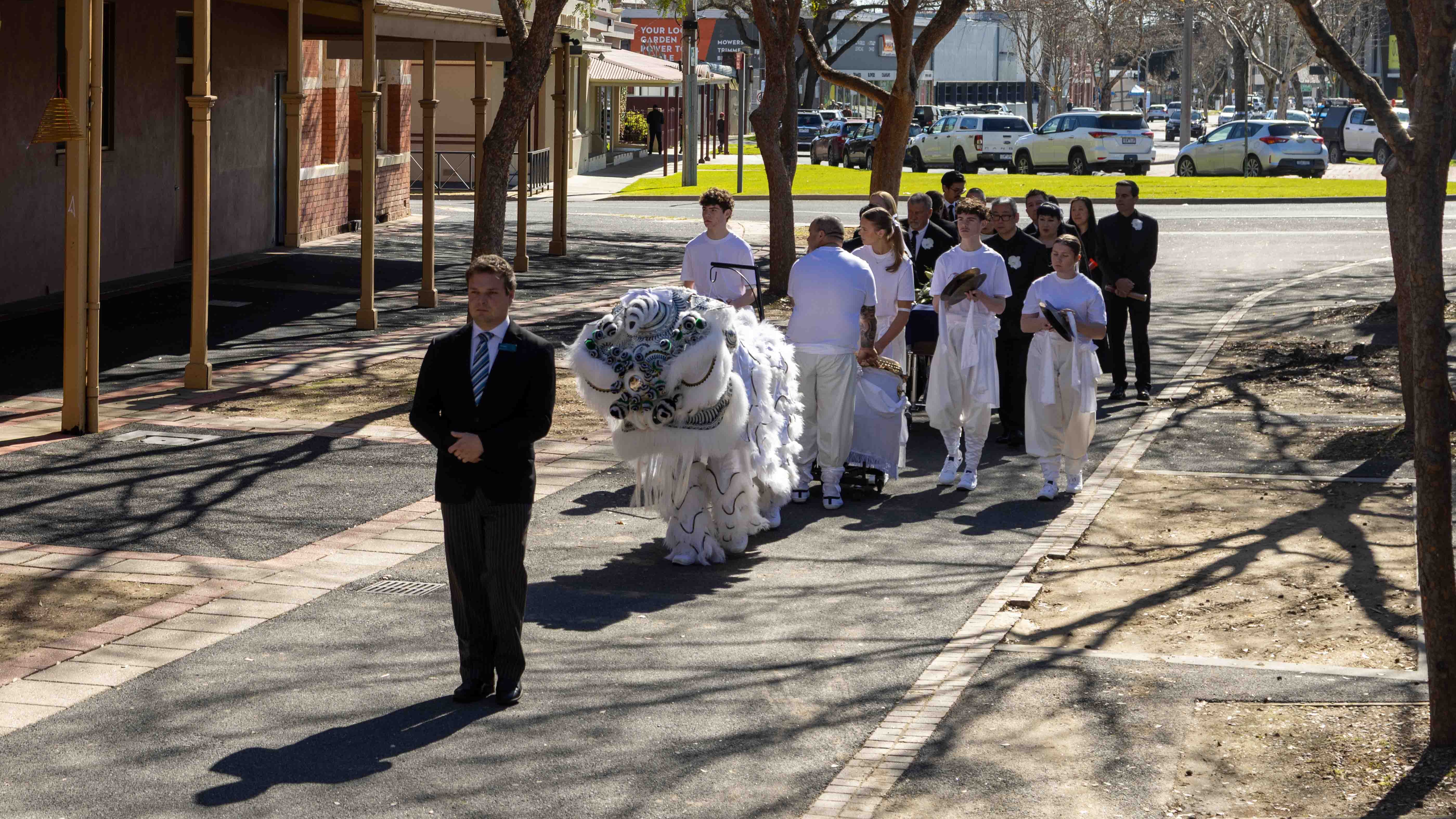 The image shows a man leading a procession of the white mourning lion, its performers, and mourners through the streets of Bendigo