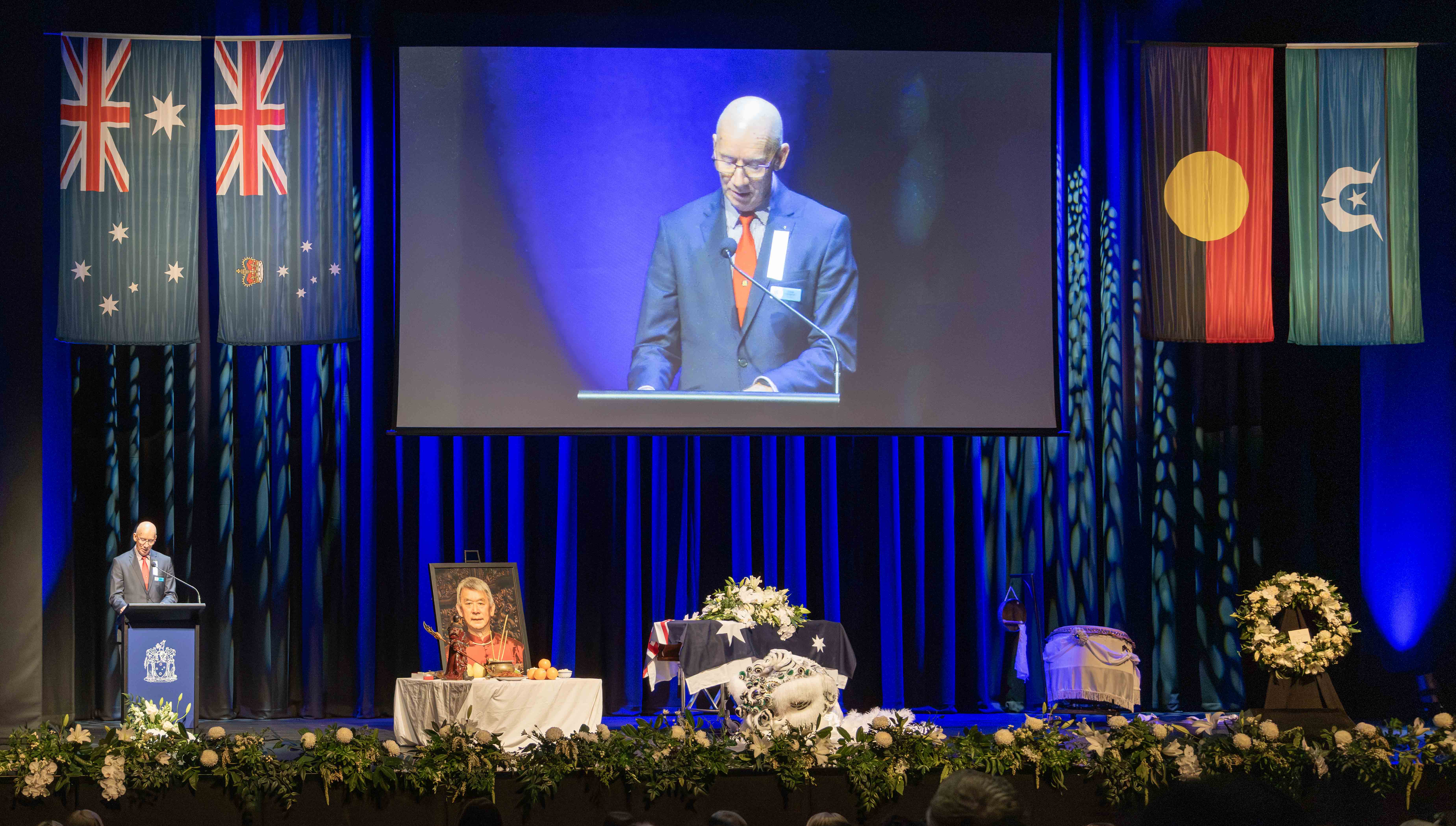 Golden Dragon Museum Chair Doug Lougoon speaks at a lectern on stage about Russell Goldfield Jack AM's contribution to Bendigo's Chinese legacy. Russell's coffin and portrait are on stage, alongside a wreath.