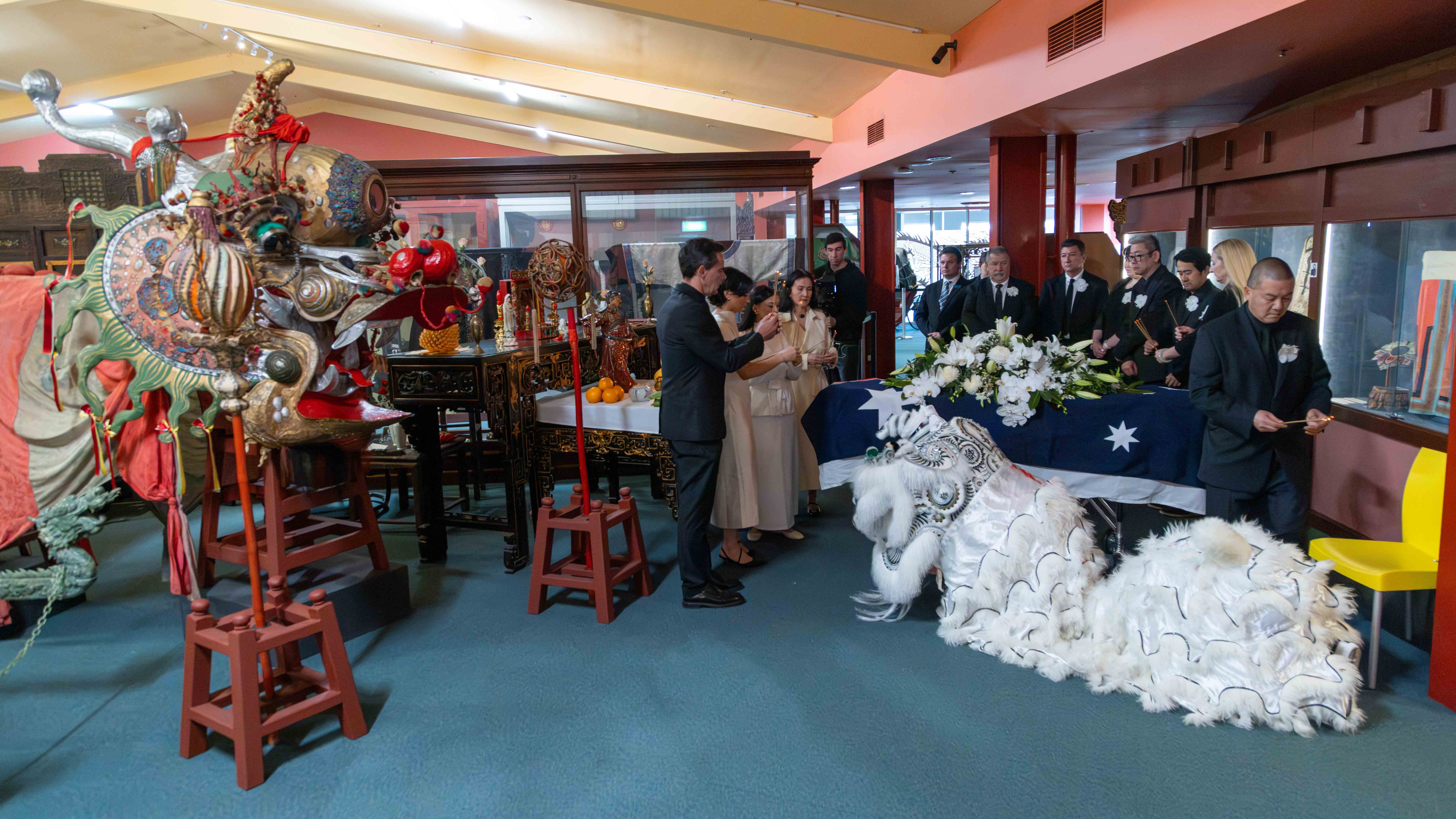 The image shows the Golden Dragon Museum's oldest dragon, Loong with the coffin of Russell Goldfield Jack AM and his white mourning lion. Russell's family stand between the two, paying tribute