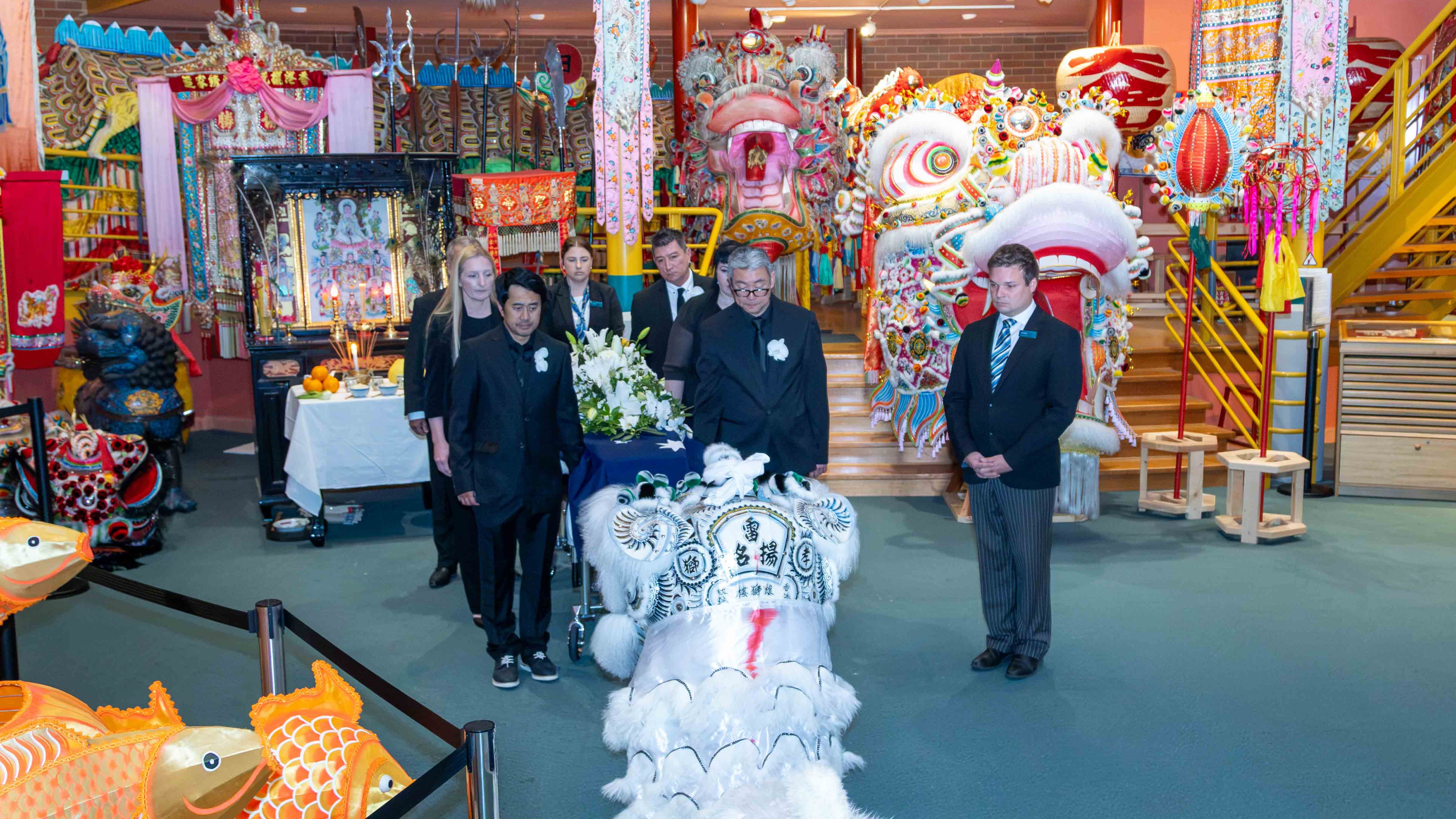 A white mourning lion accompanies Russell's coffin at the Golden Dragon Museum. It stands in front of the coffin and pallbearers, in a room filled with colourful, ceremonial lions Sun Loong and Dai Gum Loong,and other artefacts