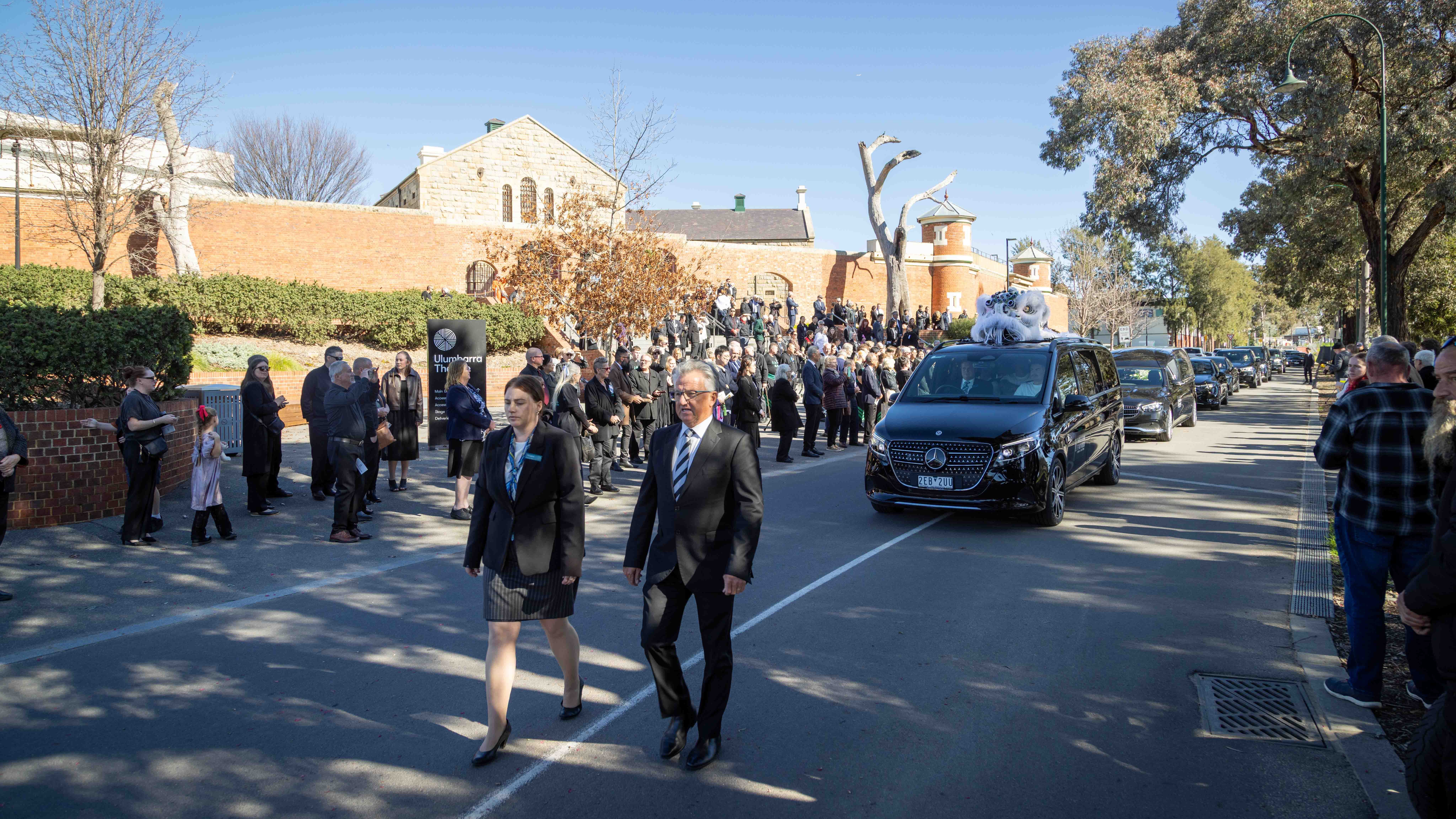 Russell's hearse leaves his State Funeral Service, accompanied by his white mourning lion, and heads to Bendigo Cemetery. Two people walk ahead of a number of cars and the road is lined with mourners