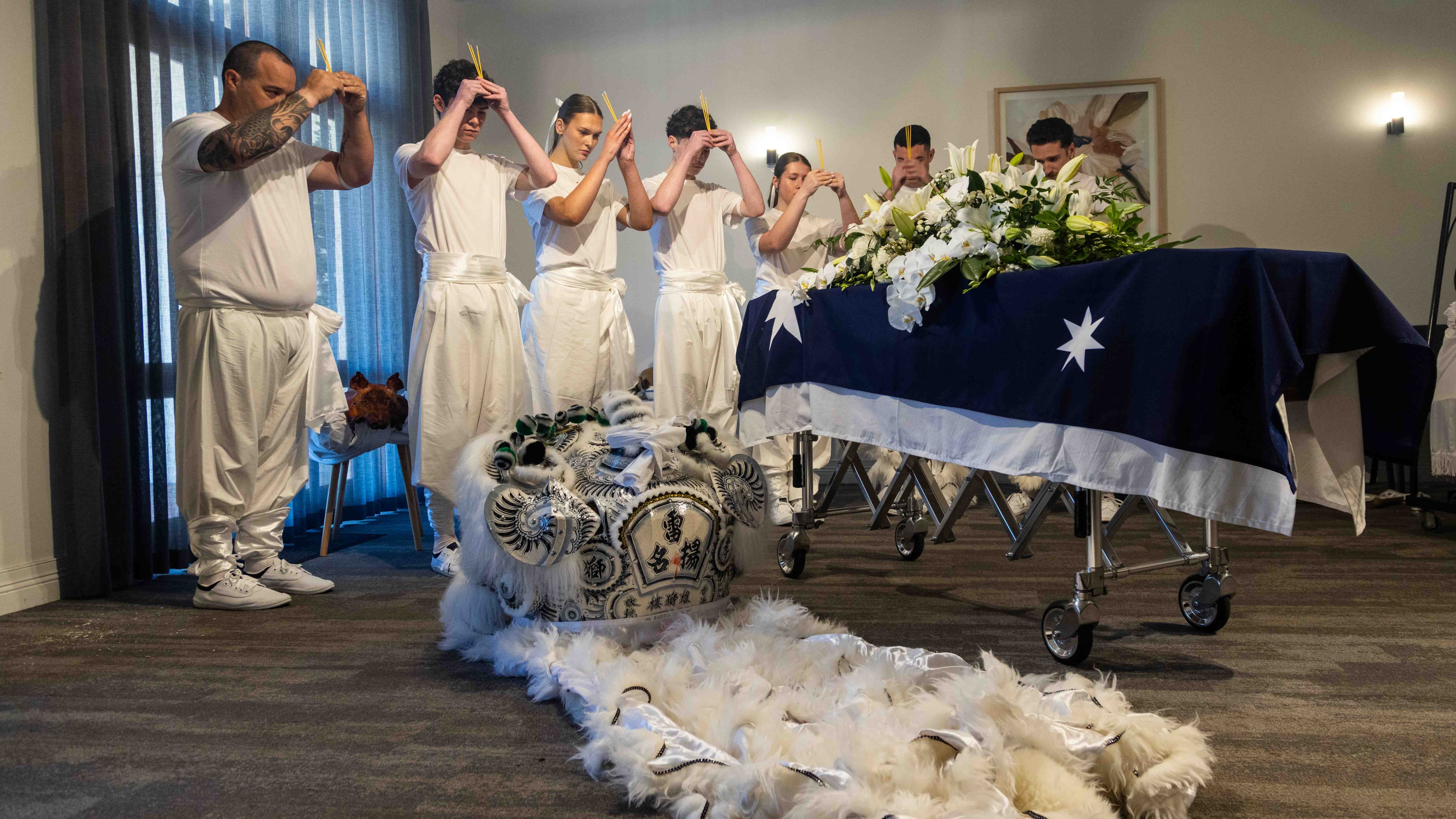 White mourning lion performers stand alongside Russell's coffin hooding joss sticks to pay tribute to him. The mourning lion is on the floor next to the coffin.