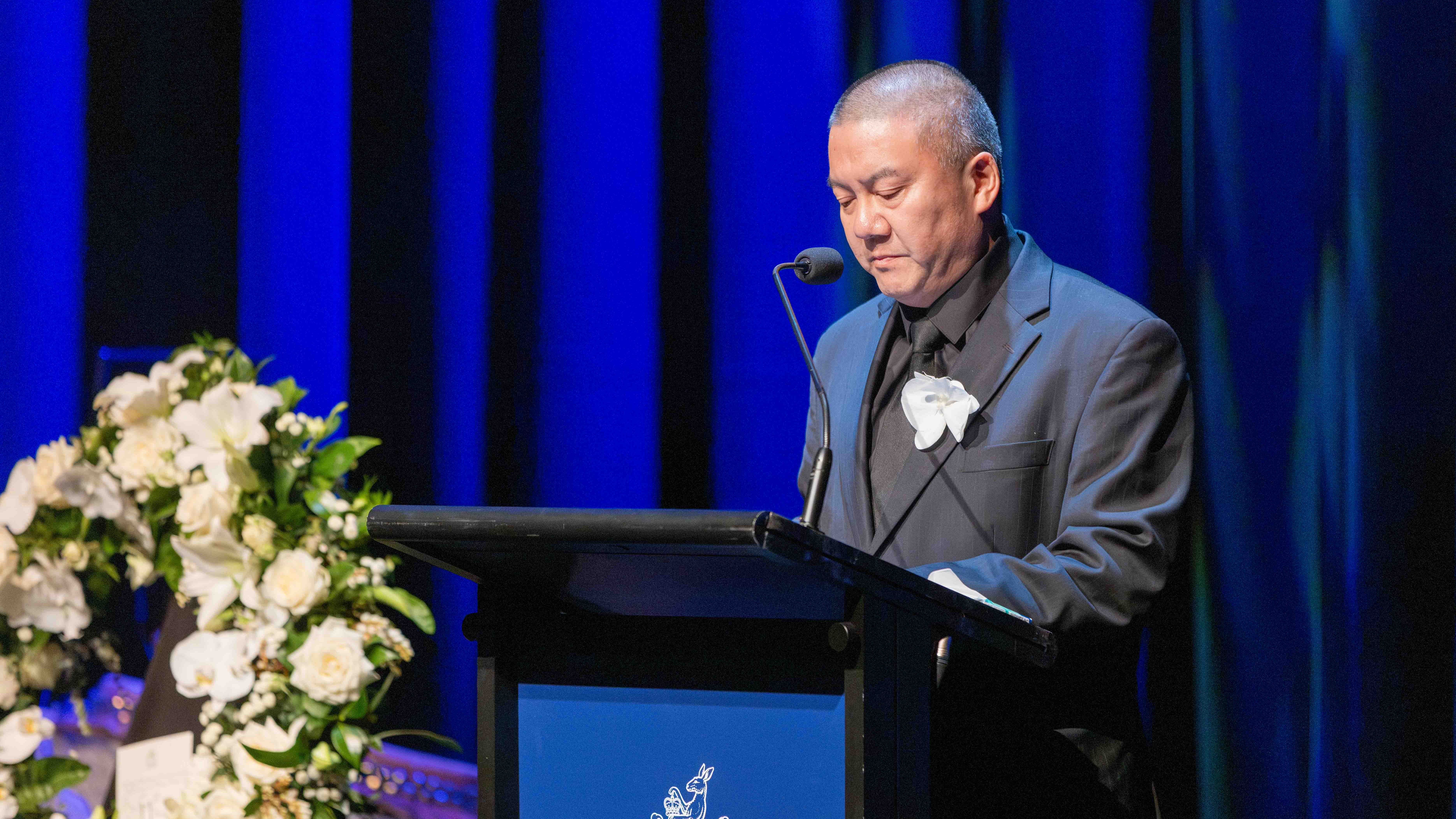 Richard Louey speaks at a lectern about Russell's connection with the Chinese community. There is a wreath to his side.