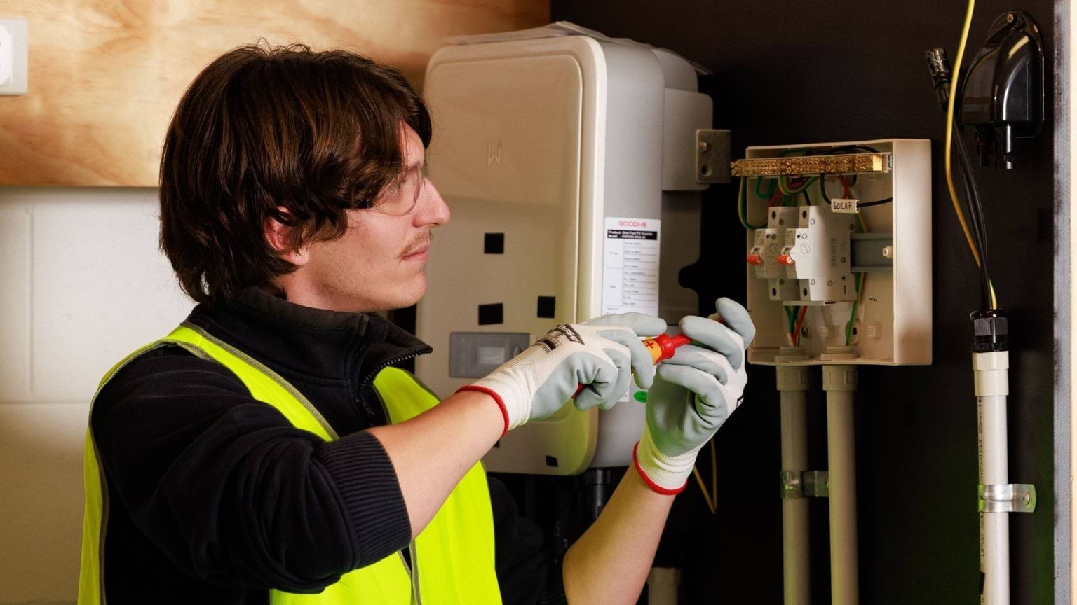A person in a yellow hi-vis vest, safety glasses and gloves uses a tool on an electrical box on a wall.