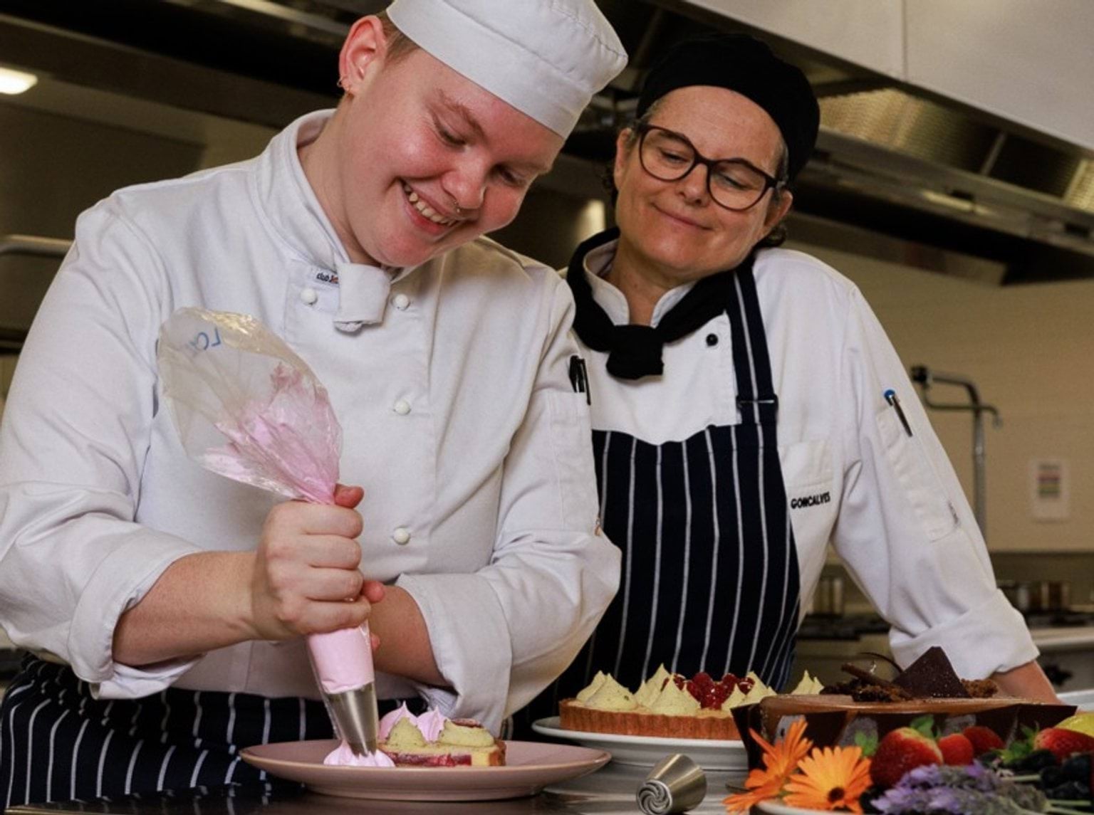 Cookery teacher smiling as she watches cookery student pipe icing onto a dessert