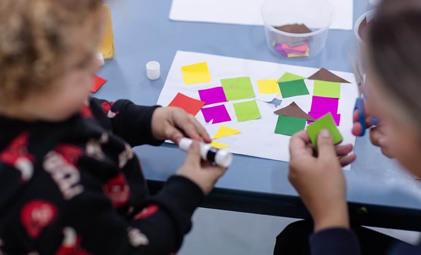 An adult cuts shapes from coloured paper while a child sticks the shapes onto a sheet of paper.