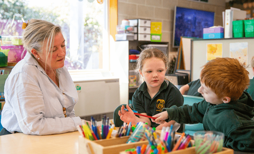 Become a teacher A female teacher talks to two children in green uniforms over a table with lots of colourful pens and pencils.